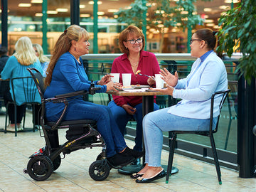 Three women sitting at a table outdoors, one using a wheelchair.